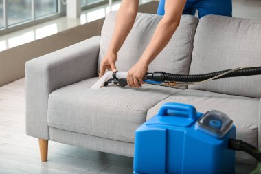 Erkek işçi, odada elektrikli süpürgeyle gri koltuktaki kiri temizliyor.