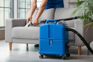 Erkek işçi, odada elektrikli süpürgeyle gri koltuktaki kiri temizliyor.