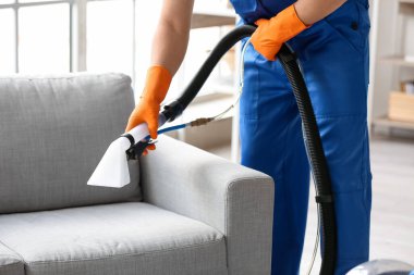 Erkek işçi, odada elektrikli süpürgeyle gri koltuktaki lekeyi temizliyor.