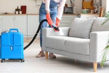 Kadın evinde elektrikli süpürgeyle gri kanepenin kirini temizliyor.