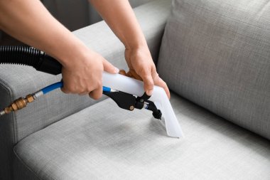 Erkek işçi, odasındaki elektrikli süpürgeyle koltuktaki kiri temizliyor.