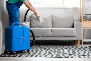 Erkek işçi, odasındaki elektrikli süpürgeyle koltuktaki kiri temizliyor.