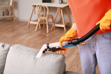 Kadın, odada elektrikli süpürgeyle gri koltuktaki kiri temizliyor.