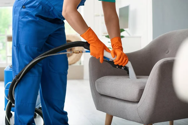 Male worker removing dirt from armchair with vacuum cleaner in room