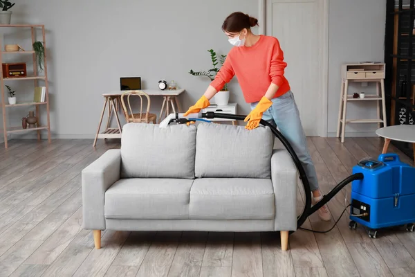 Woman removing dirt from grey sofa with vacuum cleaner in room