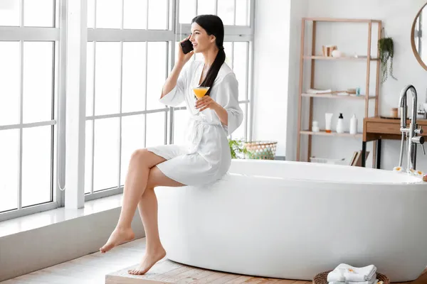 Pretty young woman with cocktail talking by mobile phone before taking bath at home