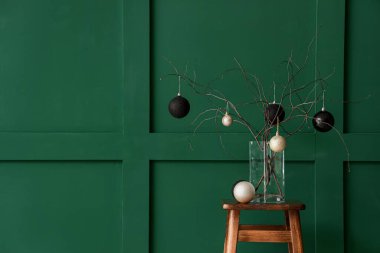 Yeşil duvarın yanındaki masada vazoda Noel topları olan ağaç dalları.
