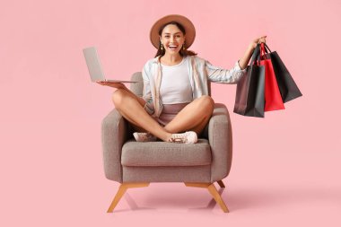 Dizüstü bilgisayarı ve alışveriş çantaları olan mutlu genç bir kadın renkli arka planda koltukta oturuyor. Kara Cuma indirimi
