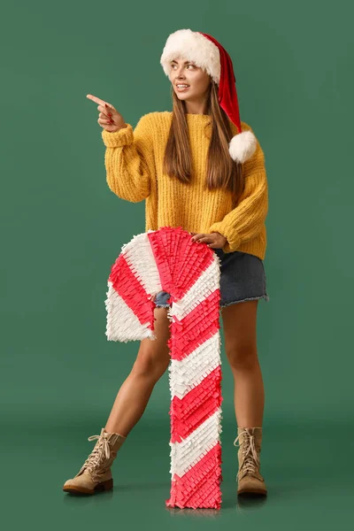 戴着圣诞礼帽 头戴棒棒糖的年轻貌美的女人指着绿色背景的东西 — 图库照片