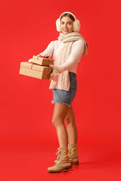 Красивая Молодая Женщина Теплых Наушниках Рождественскими Подарками Красном Фоне — стоковое фото
