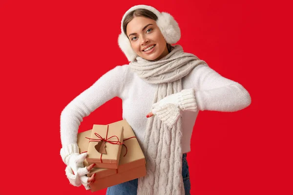 Schöne Junge Frau Warmen Ohrenschützern Zeigt Auf Weihnachtsgeschenke Auf Rotem — Stockfoto