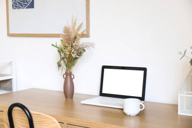 Lşık duvarının yanındaki vazoda dizüstü bilgisayar, fincan ve kurutulmuş çiçeklerle dolu bir masa.