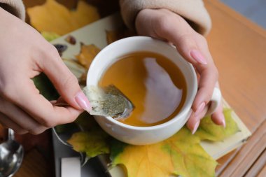 Bir fincan çay, kitap ve sonbahar yapraklı kadın elleri ahşap masada, yakın plan.