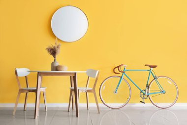 Sarı duvarın yanında vazo ve bisikletle yemek masası.