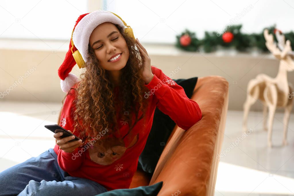 Young African-American woman in headphones with mobile phone at home on Christmas eve