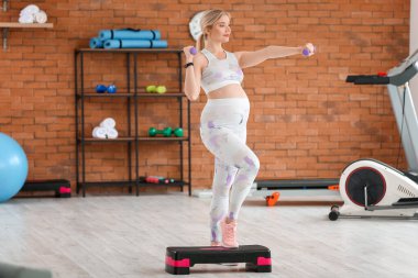 Genç hamile kadın spor salonunda dambılı ve ayakçıyla çalışıyor.