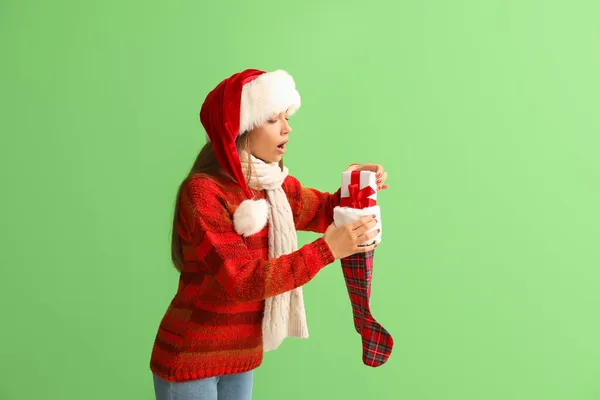令人惊讶的年轻女子从绿色背景的袜子上拿走圣诞礼物 — 图库照片