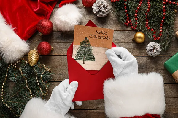 サンタクロースは封筒とギフトカードを持っていますテキストメリークリスマス木製の背景に — ストック写真