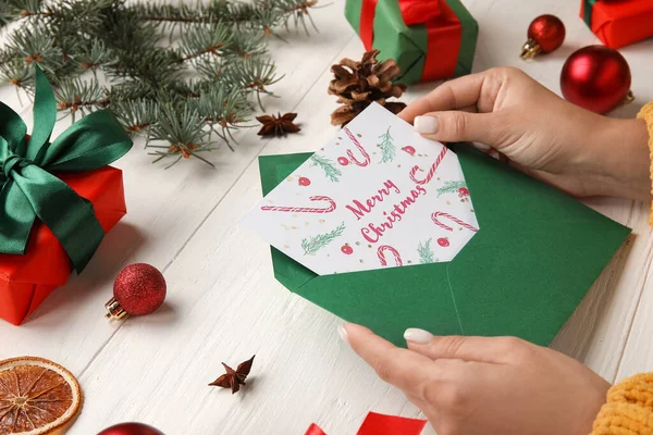 女性は白い木製のテーブルの上にテキストメリークリスマスと封筒とカードを保持 — ストック写真