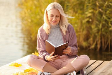Sonbahar günü nehir kenarında kitap okuyan olgun bir kadın.