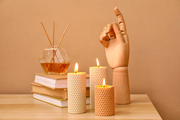 Burning candles, wooden hand, stack of books and reed diffuser on wooden table near color wall