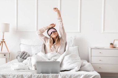 Uyku maskesi ve laptopuyla yatakta uzanan genç bir kadının sabahı.