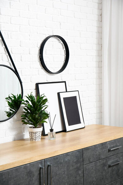 Houseplant, picture frames, reed diffuser on chest of drawers near brick wall