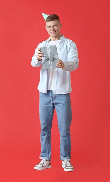 Schöner Junger Mann Mit Partyhut Und Geschenkbox Auf Rotem Hintergrund — Stockfoto