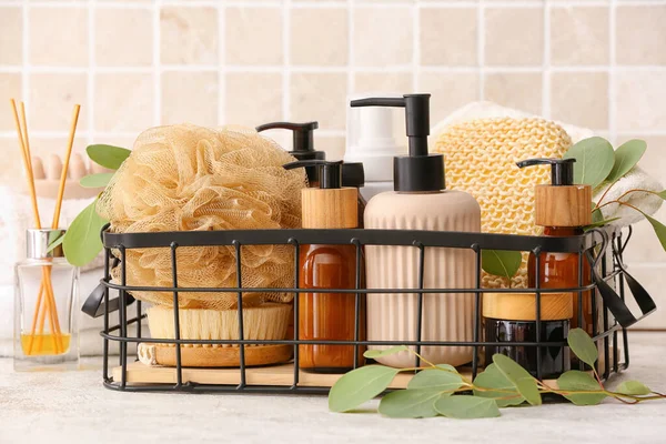 Banyo süngeri, masaj fırçası, kozmetik ürünleri şişeleri ve masa üzerinde okaliptüs dalı olan sepet.