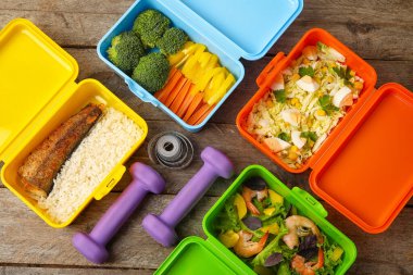 Farklı lezzetli yiyeceklere, dambıllara ve ahşap arka planda mezuralara sahip konteynırlar.