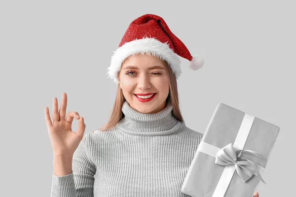 サンタの帽子の美しい若い女性とクリスマスプレゼントとともにOkジェスチャー上の灰色の背景 — ストック写真