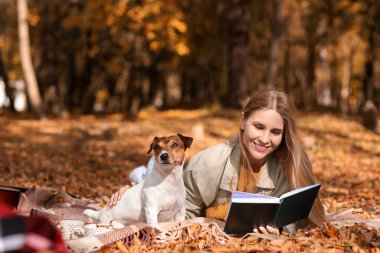 Jack Russel Terrier ile güzel bir kadın sonbahar günü parkta kitap okuyor.