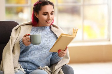 Ekoseli ve sıcak kakaolu güzel bir kadın evdeki koltukta kitap okuyor.