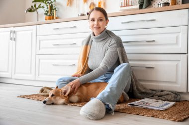 Sıcak ekoseli ve tatlı Corgi köpeği olan genç bir kadın mutfakta yerde oturuyor.