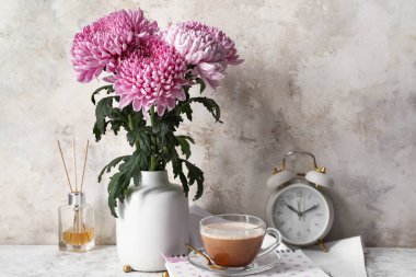 İçinde güzel kasımpatı çiçekleri, bir fincan kahve ve çalar saat olan bir vazo.