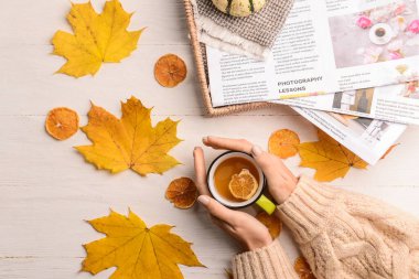 Bir fincan çay, sonbahar yaprakları ve hafif ahşap arka planda gazeteleri olan kadın elleri, yakın plan.