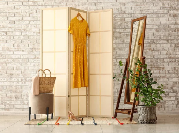 Beige folding screen with yellow dress, mirror and pouf near light brick wall