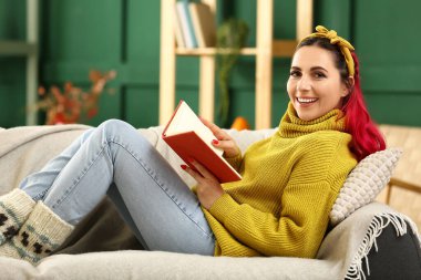 Evdeki kanepede kitap uzanmış, kalın kazaklı mutlu bir kadın.