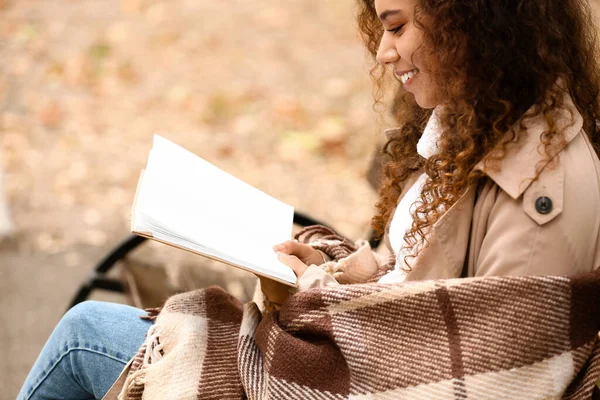 秋の公園でベンチで暖かい読書本を身に着けているアフリカ系アメリカ人女性の笑顔 — ストック写真