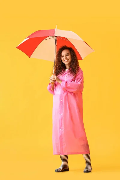若いですアフリカ系アメリカ人女性身に着けていますピンクのレインコートとガンブーツとともに傘で黄色の背景 — ストック写真