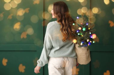 Yeşil duvarın yanında küçük bir Noel ağacı taşıyan genç bir kadın.