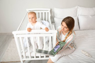 Genç anne beşikteki küçük bebeğine kitap okuyor.