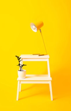 Sarı arka planda lamba ve saksı olan beyaz bir tabure.
