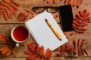 Kalemli defter, çay fincanı, cep telefonu, gözlük ve tahta arka planda bitki dalları.