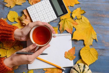 Elinde çay, defter ve sonbahar dekoru olan mavi ahşap arka planda bir kadın.