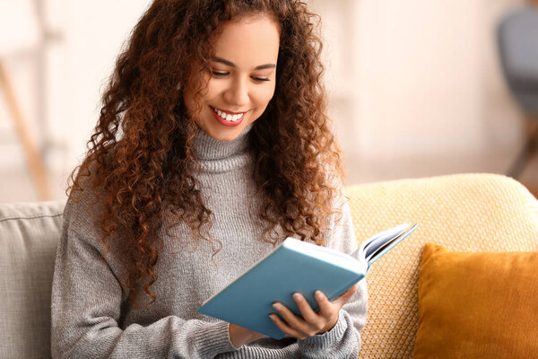 Young African-American woman reading book on sofa at home