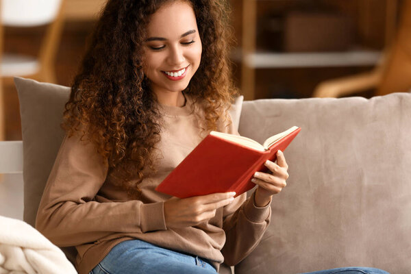 Smiling young African-American woman reading book on sofa at home