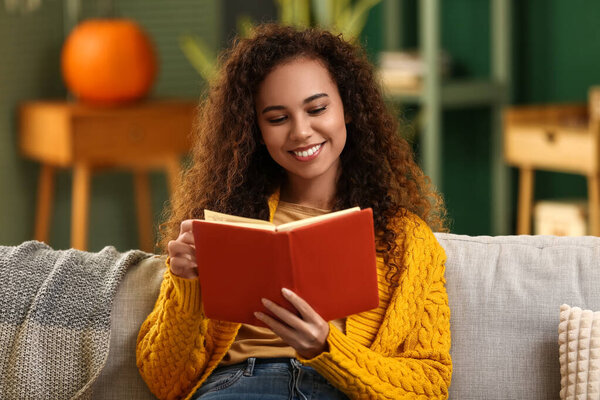 Happy young African-American woman reading book on sofa at home