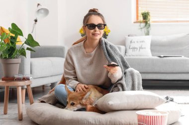 Üç boyutlu gözlüklü genç bir kadın ve evde televizyon izleyen sevimli bir Corgi köpeği.