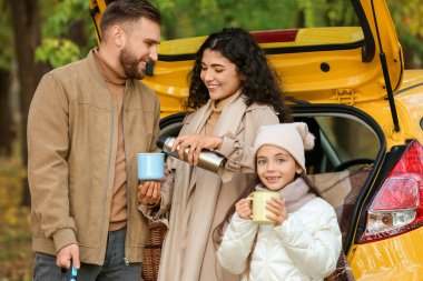 Mutlu aile, sonbahar günü sarı arabanın yanında sıcak çay içiyor.